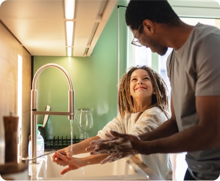 Man in front of a kitchen sink showing a child how to properly wash their hands as part of a healthy personal care routine.
